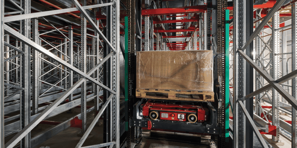 Automated storage and retrieval system (ASRS) shuttle transporting a wrapped pallet within a high-density warehouse racking structure, illustrating material handling under peak seasonal demand conditions